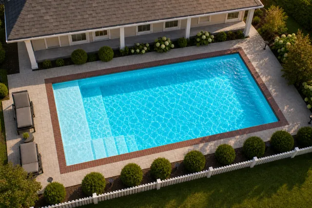 Daytime render of the The Vision 16 fiberglass pool with brick coping and a Coral Blue interior, seen from above at an angle.