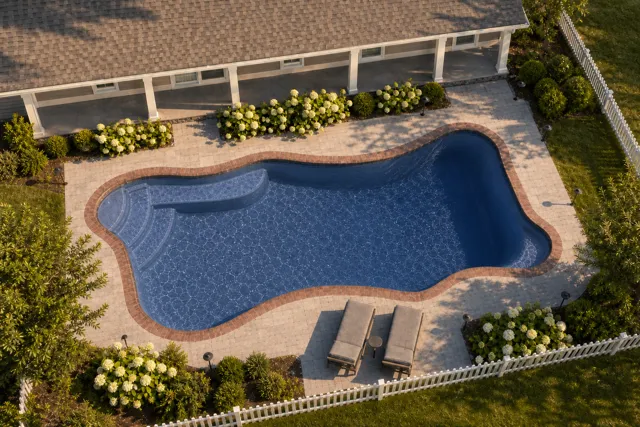 Daytime render of the Cape Cod 12 fiberglass pool with brick coping and a Ocean Blue interior, seen from above at an angle.